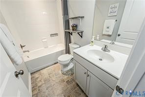 Bathroom featuring a single-sink vanity with white countertop, chrome faucet, tiled shower-tub surround, dual wall-mounted shelving, and stone-finish flooring