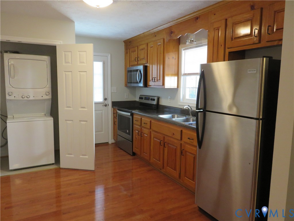 Kitchen featuring wooden cabinetry, stainless steel appliances, dark countertops, and wood-finish flooring, complemented by a dedicated laundry area Kitchen featuring wooden cabinetry, stainless steel appliances, dark countertops, and wood-finish flooring, complemented by a dedicated laundry area