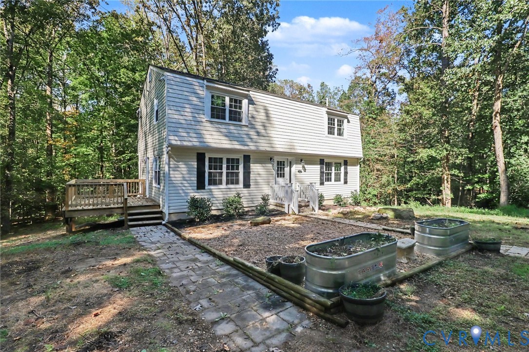 Two-story residence with horizontal siding, a raised wood deck, stone paver pathway, and galvanized steel garden beds situated on a wooded lot