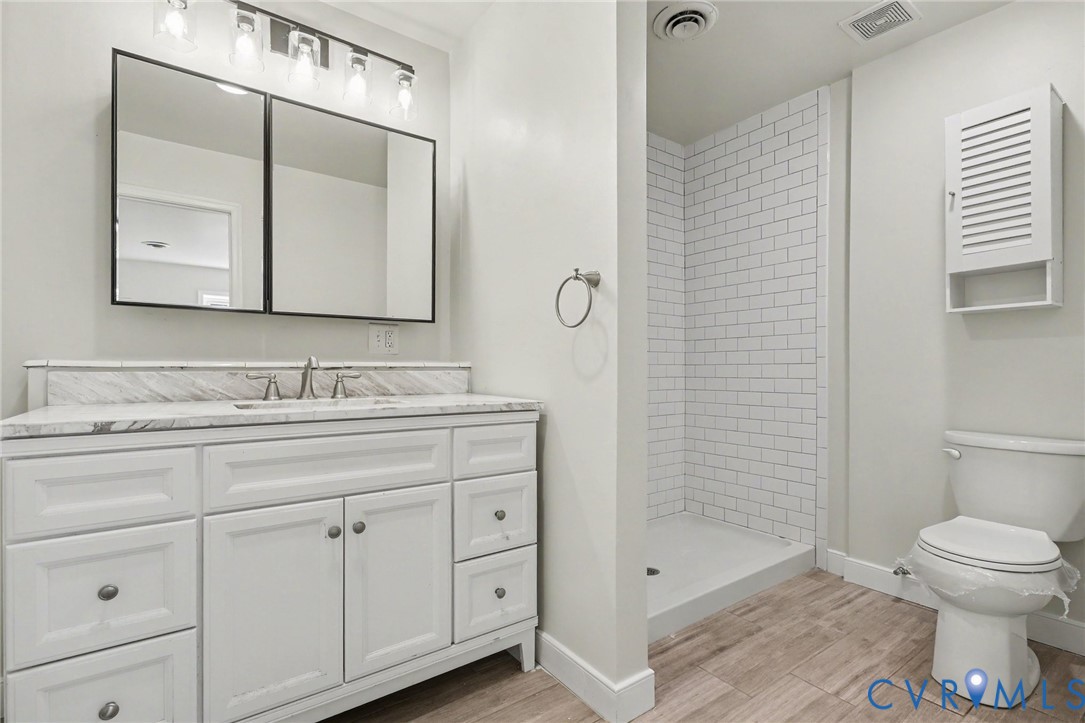 White shaker-style vanity with stone-top surface, dual-panel medicine cabinet, walk-in shower with white subway tile surround, and wood-finish flooring