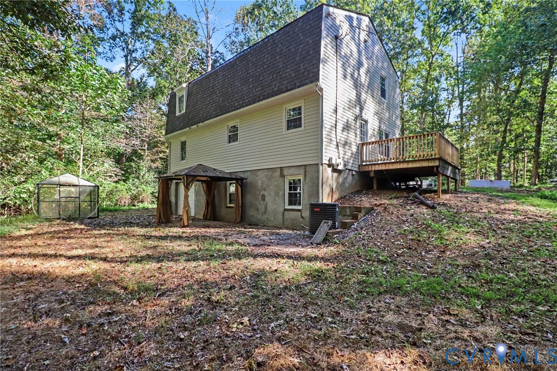 Multi-story residence featuring light-toned vinyl siding, a shingled gambrel roof, an elevated wood deck, and a ground-level covered patio area set within a wooded lot