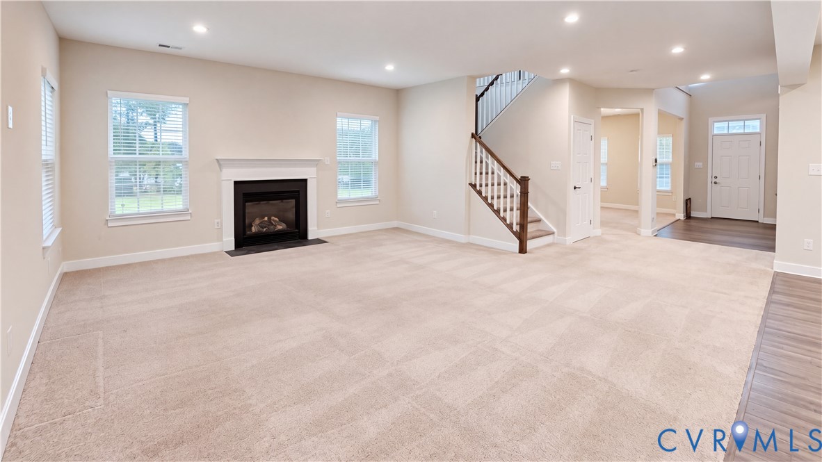 Open-concept interior featuring a fireplace with a white mantel, recessed lighting, carpeted flooring, and a staircase with a wooden handrail Open-concept interior featuring a fireplace with a white mantel, recessed lighting, carpeted flooring, and a staircase with a wooden handrail