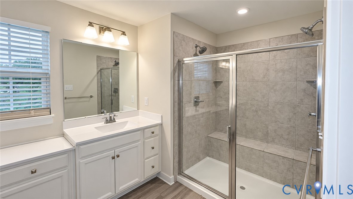 Bathroom featuring a walk-in shower with tile surround and built-in bench, vanity with white countertop, integrated sink, and wood-finish flooring