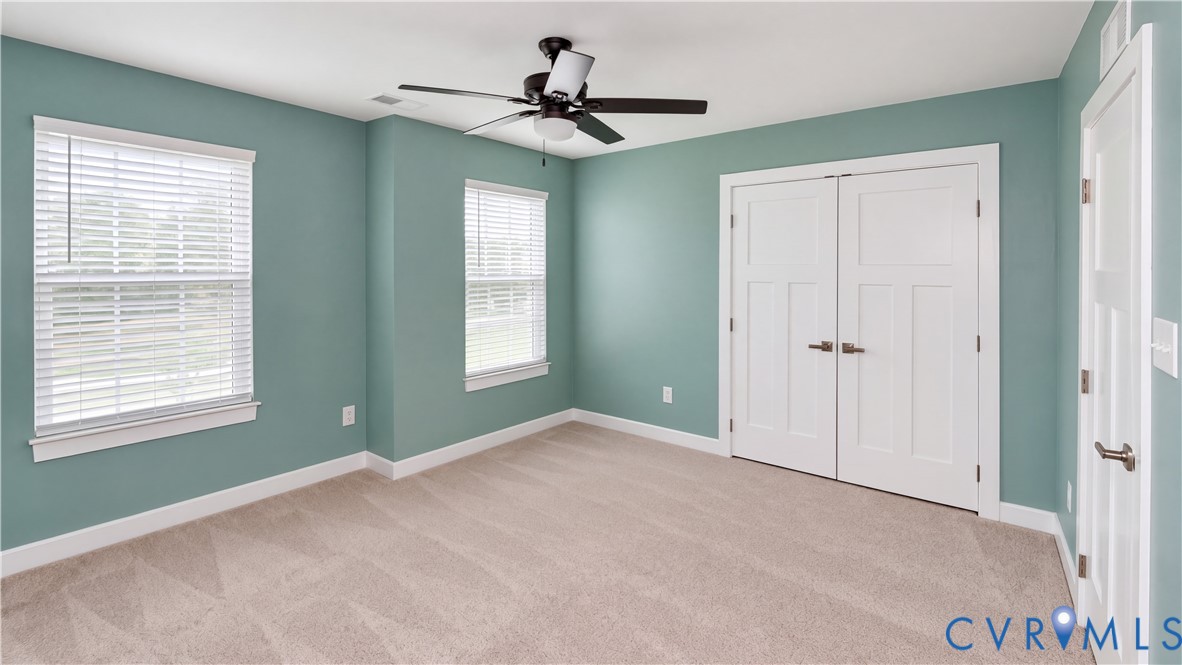 Neutral-toned carpeting, dual-window configuration with horizontal blinds, central ceiling fan with integrated lighting, and white shaker-style double closet doors