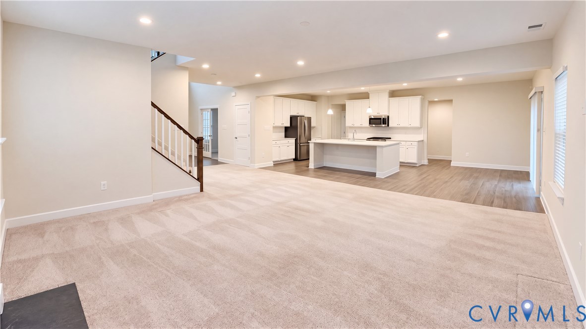 Open-concept interior featuring a kitchen with white cabinetry and a center island, wood-finish flooring, recessed lighting, and a staircase with a wooden handrail Open-concept interior featuring a kitchen with white cabinetry and a center island, wood-finish flooring, recessed lighting, and a staircase with a wooden handrail