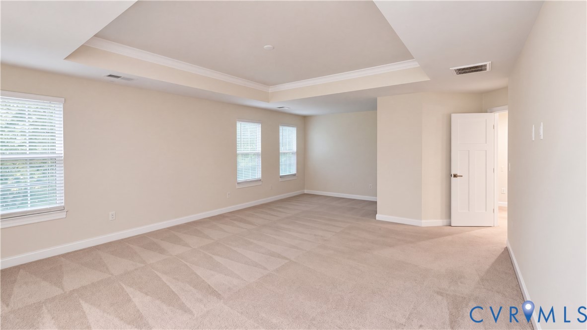 Expansive room featuring a tray ceiling, neutral wall-to-wall carpeting, multiple windows with blinds, and a multi-panel interior door Expansive room featuring a tray ceiling, neutral wall-to-wall carpeting, multiple windows with blinds, and a multi-panel interior door
