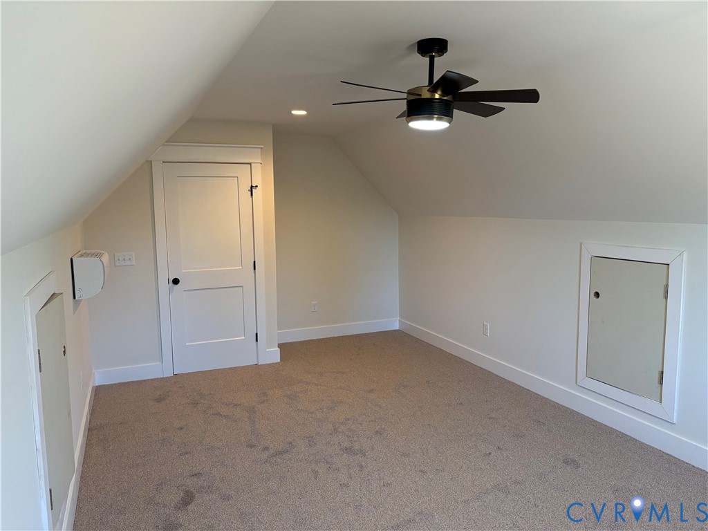 Finished attic space featuring vaulted ceilings, neutral wall-to-wall carpeting, a multi-blade ceiling fan with integrated lighting, and two built-in access doors