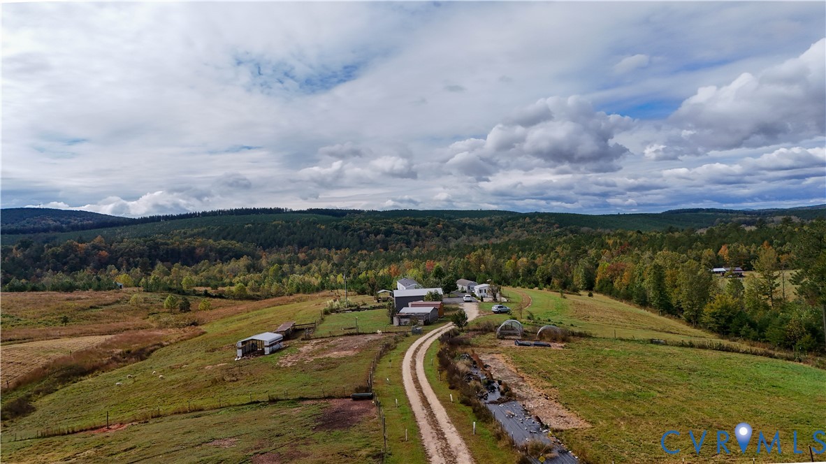 Expansive acreage featuring rolling grassy fields, multiple outbuildings, a winding gravel driveway, and a natural creek, set against a backdrop of mature woodland and distant mountain ranges