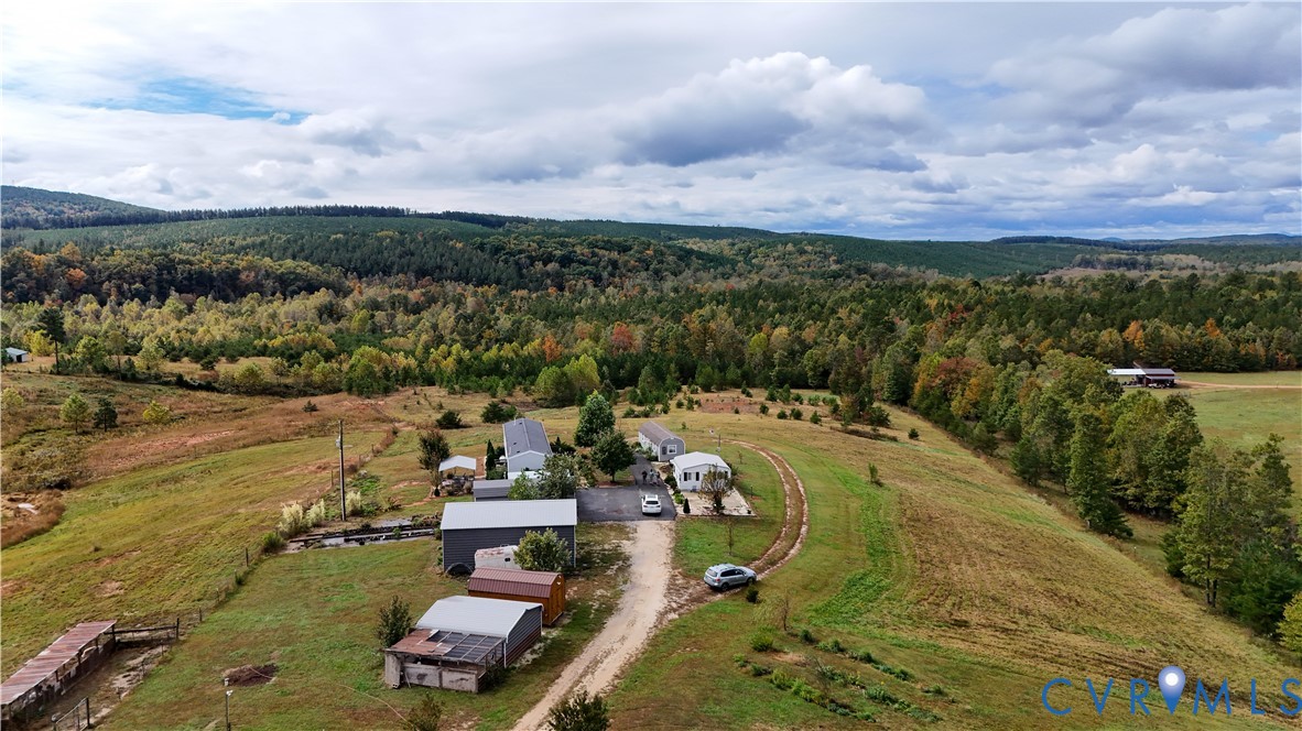 Expansive acreage featuring multiple outbuildings, open pasture land, and a gravel driveway set against a forested mountain backdrop