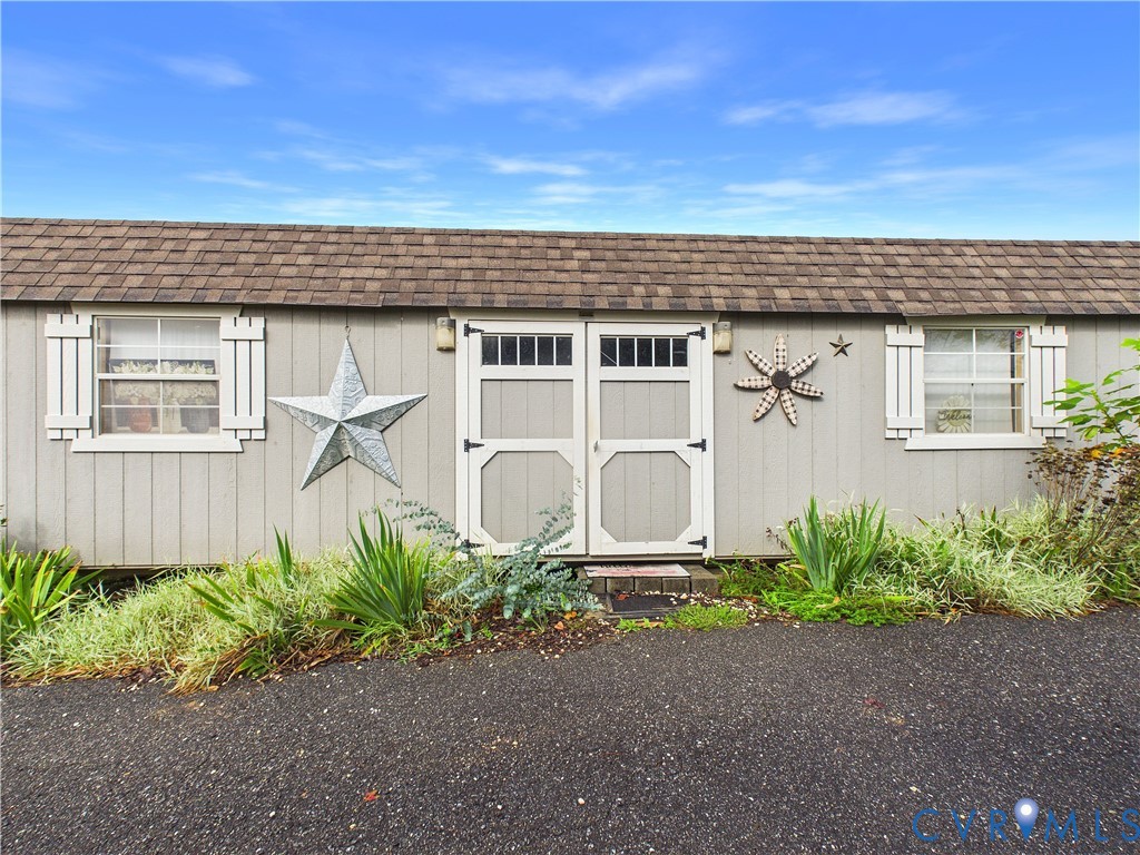 Storage structure featuring vertical panel siding, double doors with transom windows, dual windows with shutters, and asphalt shingle roofing