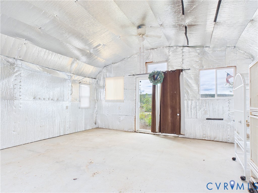 Enclosed structure featuring reflective foil insulation, vaulted ceiling with a central ceiling fan, concrete flooring, multiple windows, and a sliding glass door