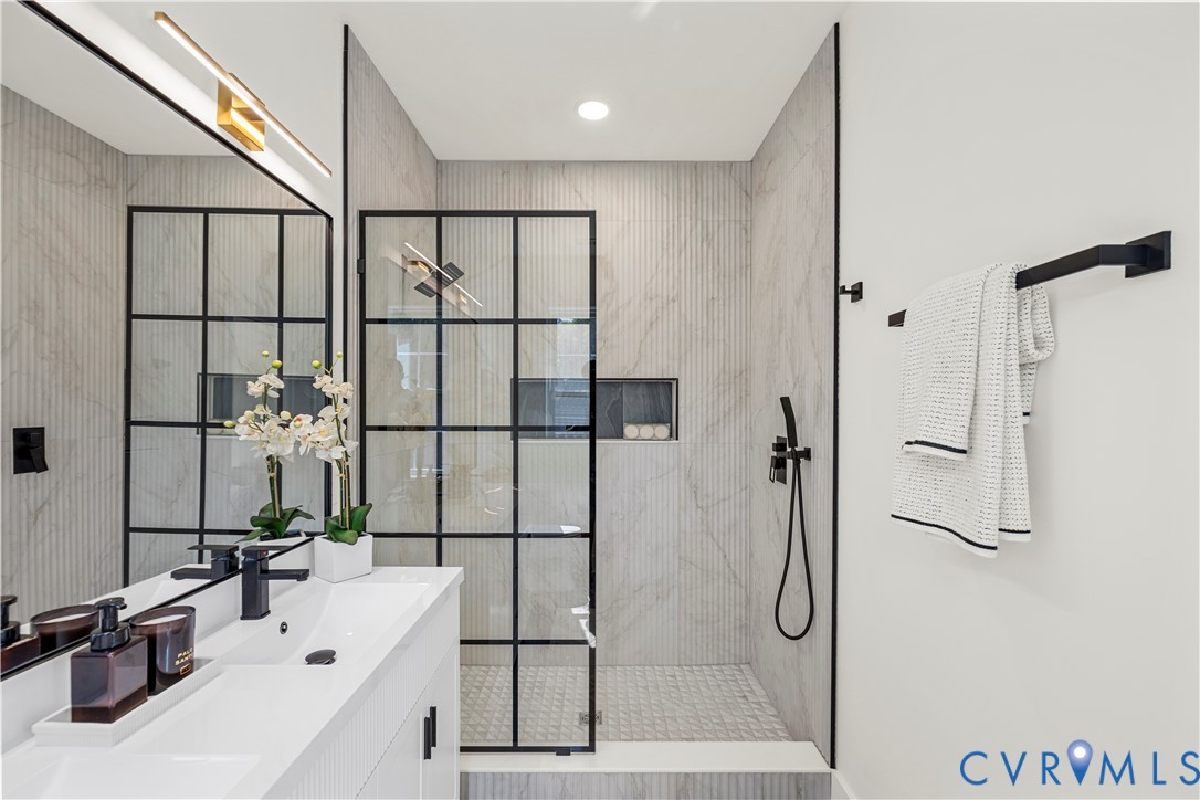 Walk-in shower featuring a grid-patterned black frame glass enclosure, floor-to-ceiling textured tile wall, recessed niche, and matte black handheld shower fixture, complemented by a dual-sink vanity with matte black hardware