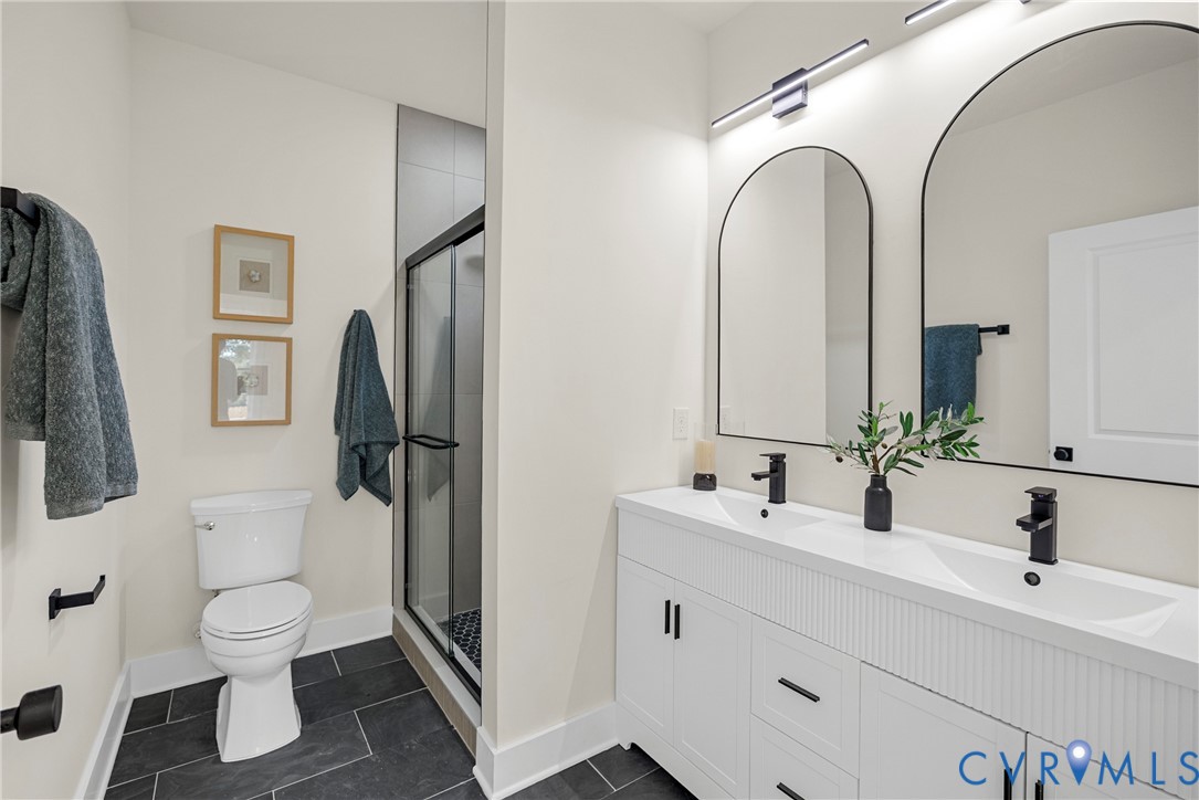 Double vanity with integrated sinks, matte black fixtures, arched vanity mirrors, walk-in glass-enclosed shower, and dark tile flooring