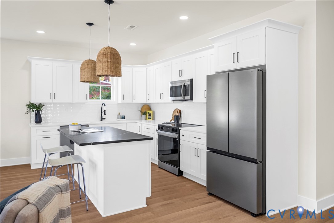 Kitchen featuring white shaker-style cabinetry, stainless steel appliances, dark stone-finish island countertop, woven pendant lighting, and wood-finish flooring