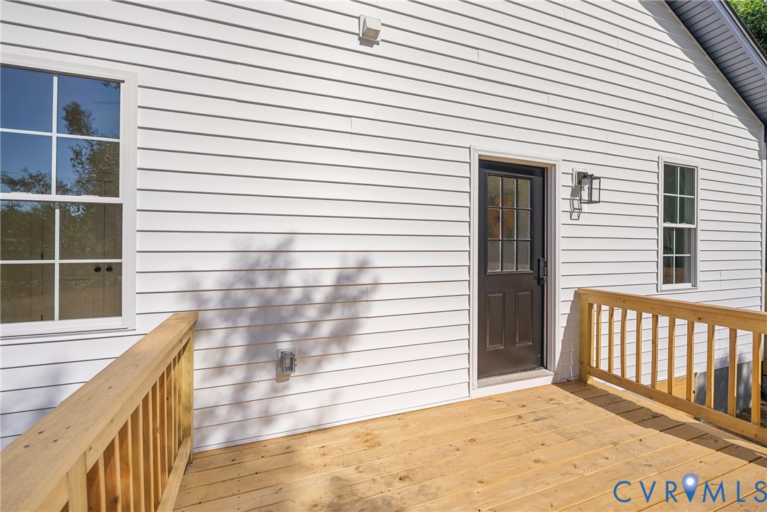 Exterior deck with wood-finish decking and wooden railing, featuring white horizontal siding, double-hung windows with muntins, and a dark-toned door with a glass inlay