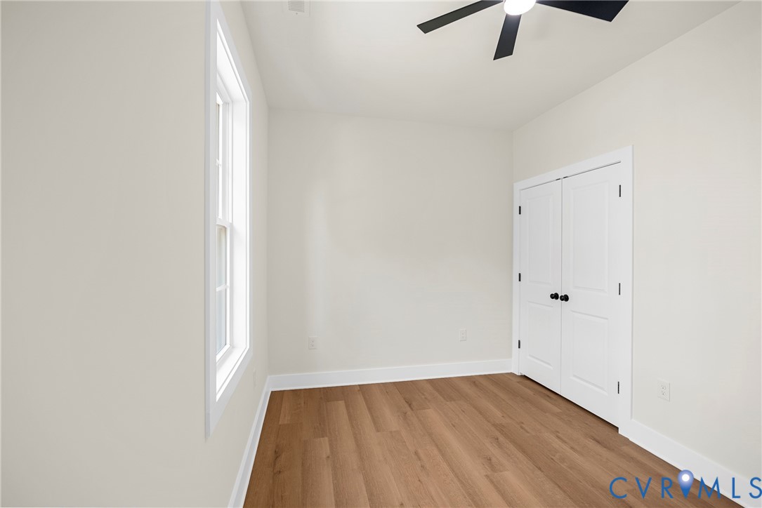 Neutral-toned interior featuring wood-finish flooring, a double-door closet with matte black hardware, a ceiling fan with integrated lighting, and a single-pane window