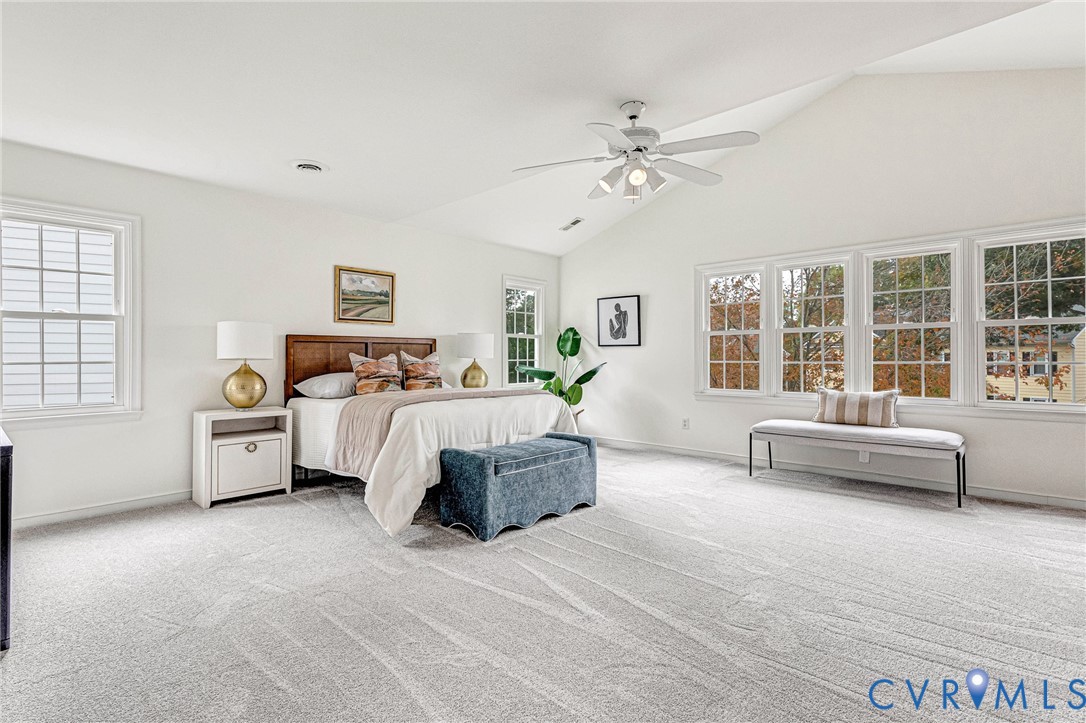 Spacious bedroom featuring vaulted ceilings, neutral wall-to-wall carpeting, a multi-pane window configuration, and a ceiling fan with integrated lighting