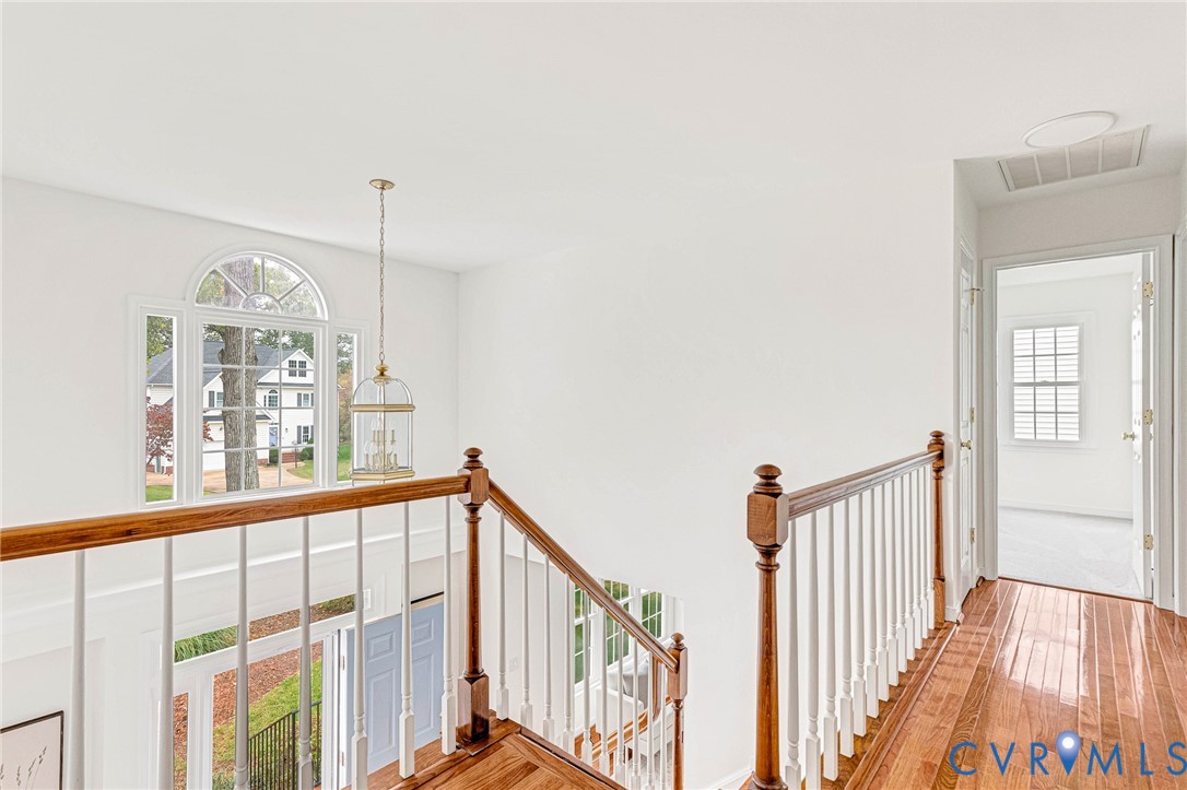 Landing featuring wood-finish flooring, a traditional staircase with a wooden handrail, white spindle balusters, a decorative hanging pendant light, and an arched window