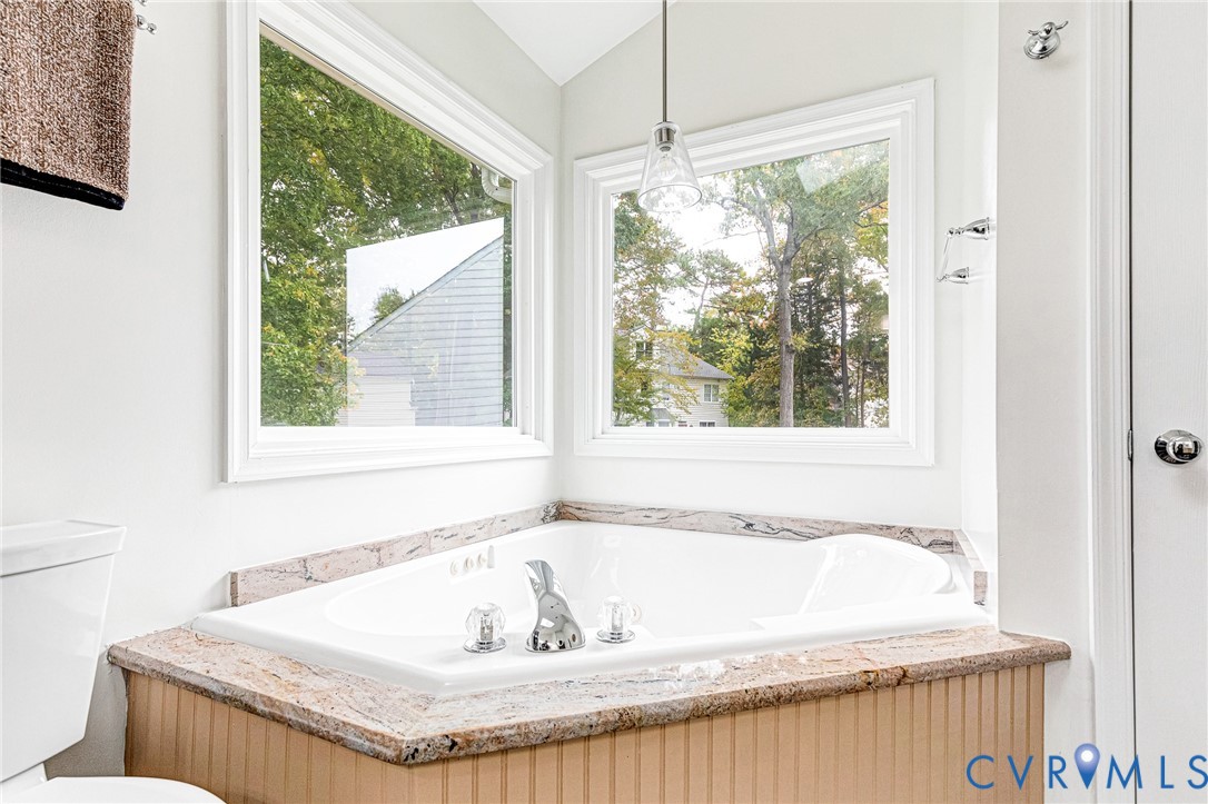 Corner soaking tub featuring a stone-surface surround, beadboard paneling, chrome fixtures, and dual picture windows with pendant lighting