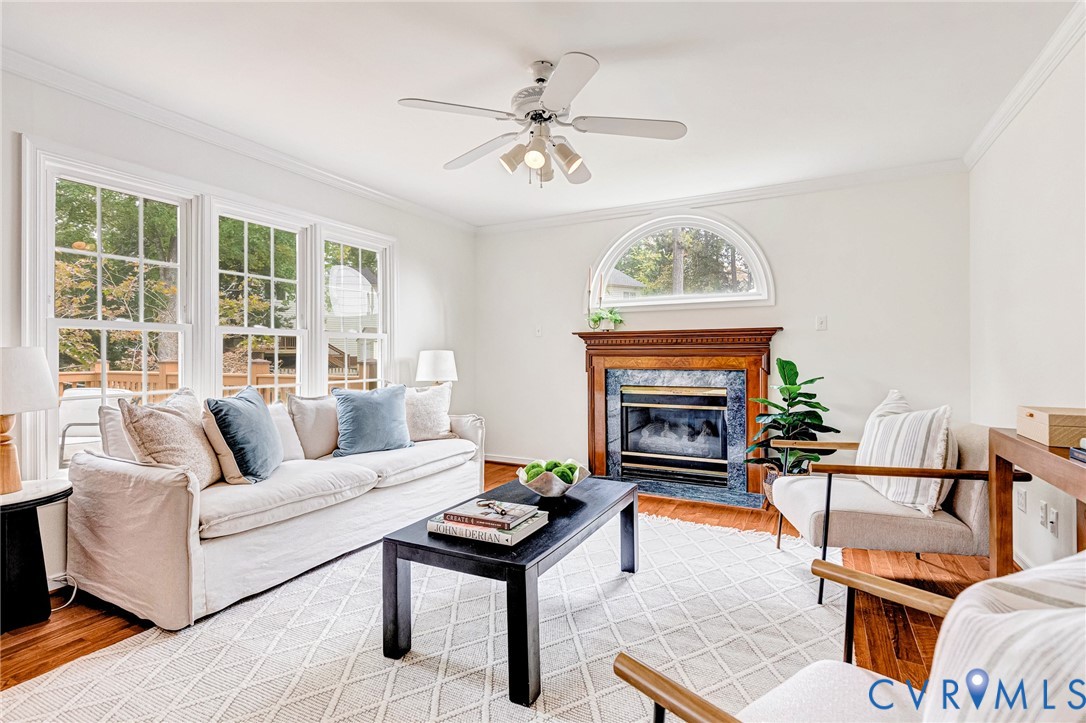 Living area featuring wood-finish flooring, a fireplace with a decorative mantel and stone surround, an arched window, crown molding, and a multi-light ceiling fan