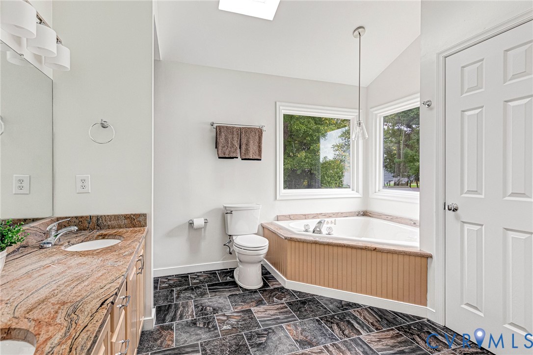 Stone countertop vanity, large soaking tub with tile surround, vaulted ceiling with skylight, and stone-finish floor tile