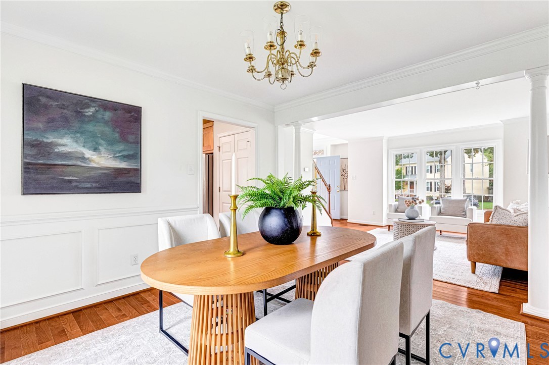 Dining area featuring wood-finish flooring, wainscoting, an overhead brass chandelier, and white structural columns Dining area featuring wood-finish flooring, wainscoting, an overhead brass chandelier, and white structural columns