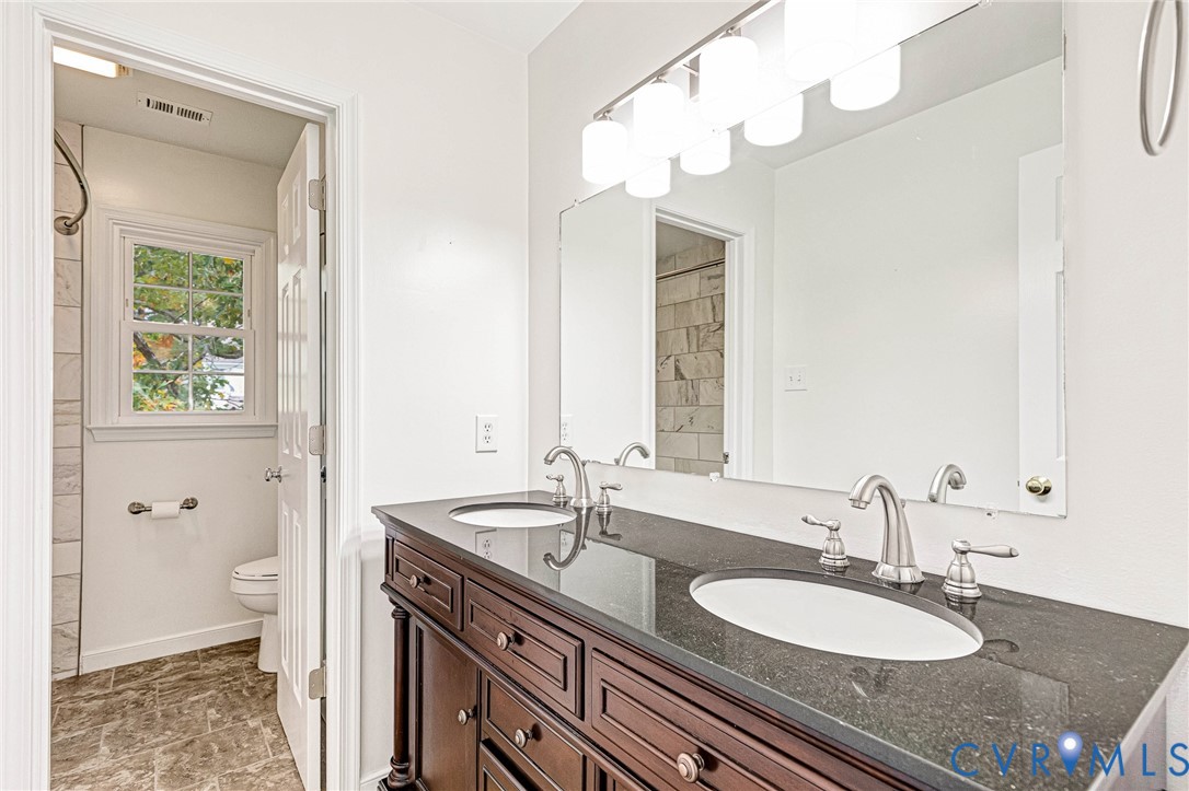 Double vanity featuring a dark stone countertop, dual undermount sinks, wood-finish cabinetry, a large wall-mounted mirror, and multi-bulb vanity lighting
