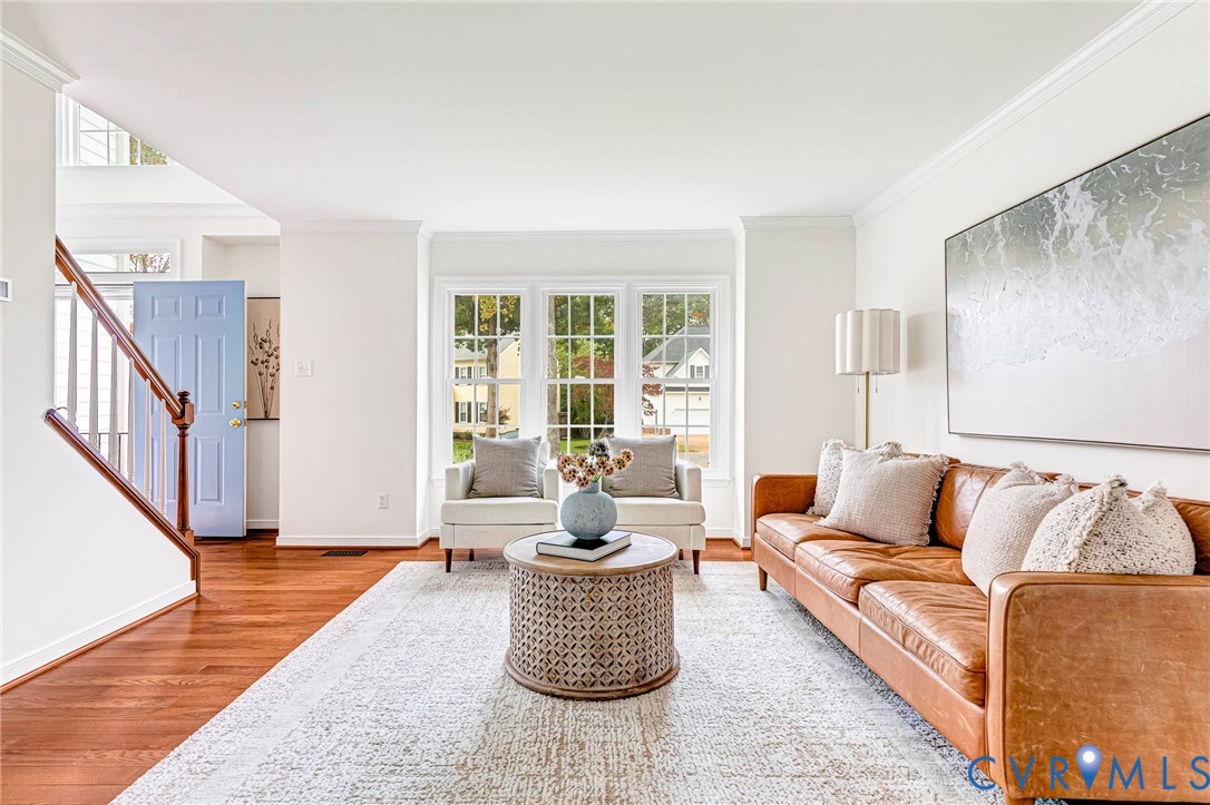 Living space featuring wood-finish flooring, a triple-pane window unit, crown molding, and a staircase with a wooden handrail and balusters Living space featuring wood-finish flooring, a triple-pane window unit, crown molding, and a staircase with a wooden handrail and balusters