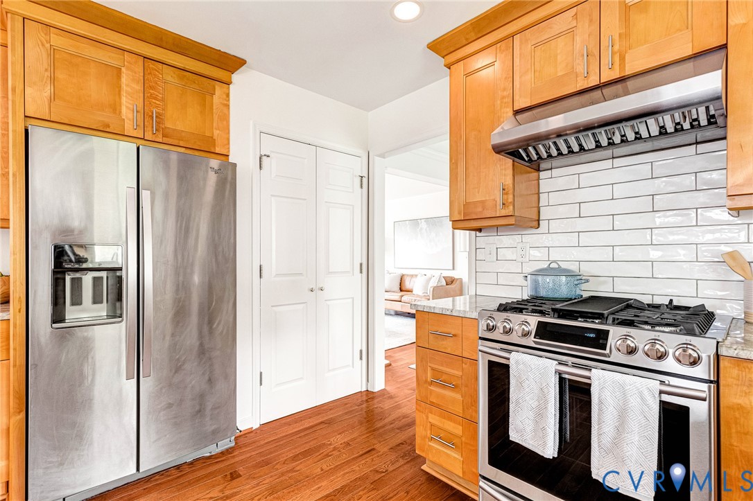 Kitchen featuring custom wood cabinetry, stainless steel appliances, a professional-grade gas range with a matching range hood, a white subway tile backsplash, and wood-finish flooring
