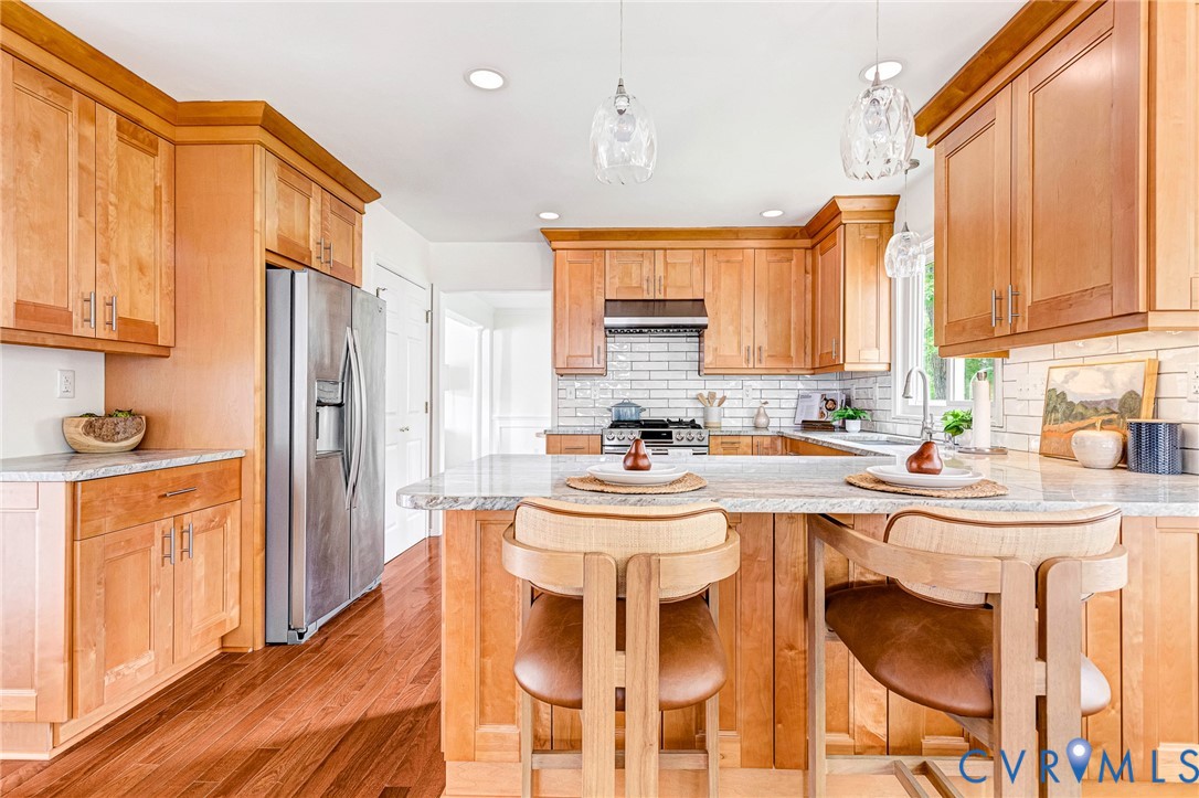 Kitchen featuring natural-finish wood cabinetry, stone countertops, stainless steel appliances, subway tile backsplash, and overhead glass pendant lighting