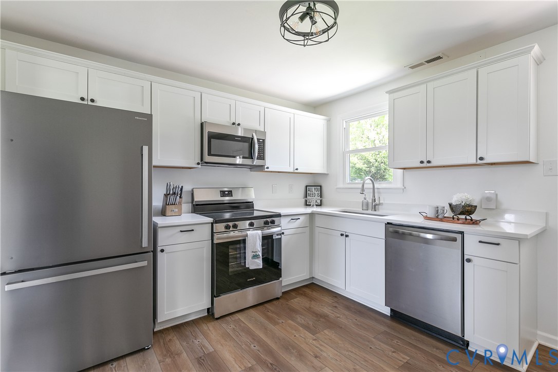 Kitchen featuring white shaker-style cabinetry, stainless steel appliances, solid surface countertops, wood-finish flooring, and a modern drum-style flush-mount light fixture