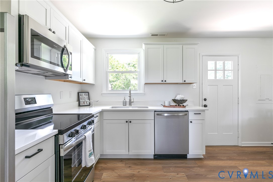 Kitchen featuring white Shaker-style cabinetry, stainless steel appliances, wood-finish flooring, solid-surface countertops, and a six-pane glass inset door