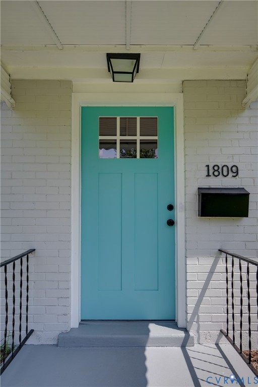 Painted brick exterior entry featuring a paneled door with a six-pane window, black metal hardware, a wall-mounted mailbox, and a recessed ceiling light fixture