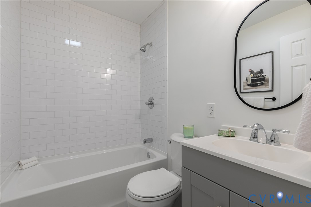 Bathroom featuring white subway tile tub surround, grey shaker-style vanity, solid surface countertop with integrated sink, chrome hardware, and an oval mirror with a black frame