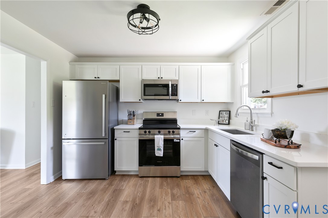 Kitchen featuring white shaker-style cabinetry, stainless steel appliances, white solid-surface countertops, wood-finish flooring, and a matte black geometric flush-mount light fixture