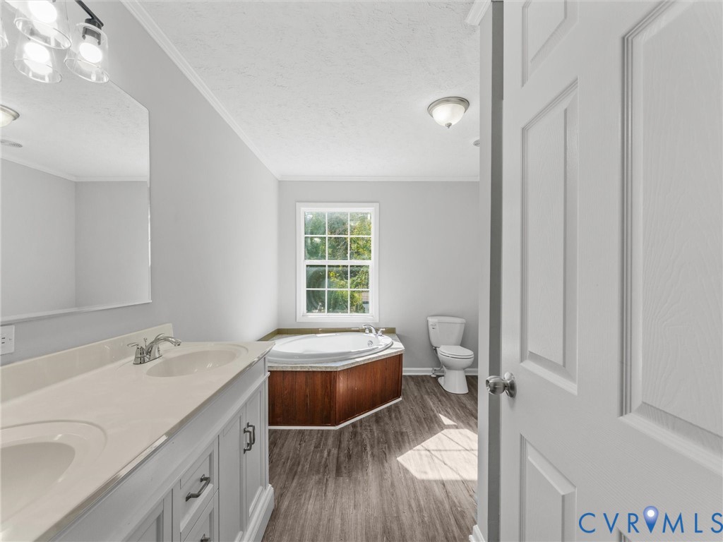 Dual-sink vanity with a solid-surface countertop, jetted soaking tub with wood-finish surround, wood-finish flooring, and multi-light wall fixtures