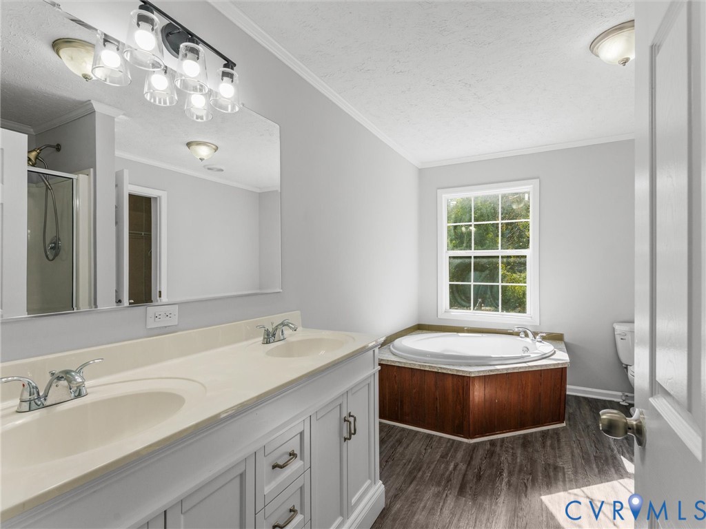 Dual-sink vanity with light-toned countertop, large wall-mounted mirror, multi-light fixture, circular jetted soaking tub with wood-finish surround, and wood-finish flooring