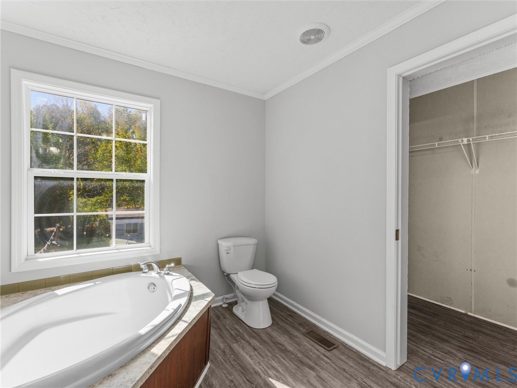 Oval soaking tub with stone-composite surround, multi-pane window, wood-finish flooring, and walk-in closet with built-in shelving