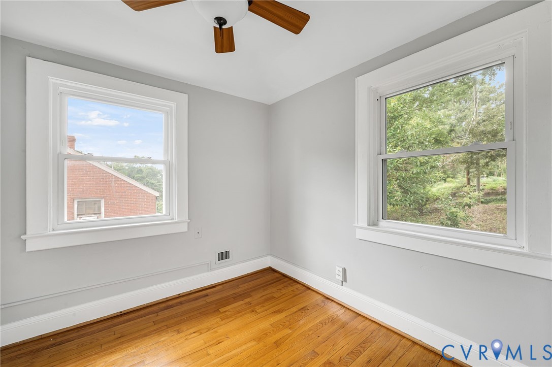 Neutral-toned interior room featuring wood-finish flooring, two double-hung windows with white casing, and a ceiling-mounted fan with wooden blades