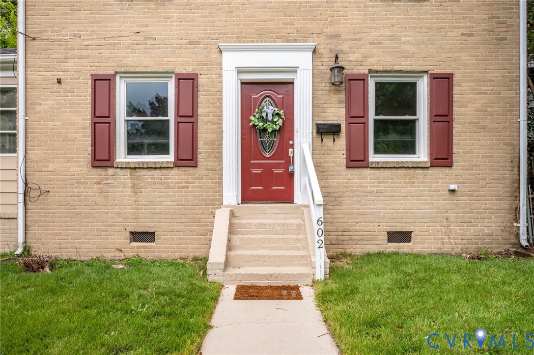 Exterior facade featuring tan brick masonry, red shutters, a central entrance with a white fluted door surround, an oval glass-paneled door, and concrete steps with a white handrail Exterior facade featuring tan brick masonry, red shutters, a central entrance with a white fluted door surround, an oval glass-paneled door, and concrete steps with a white handrail