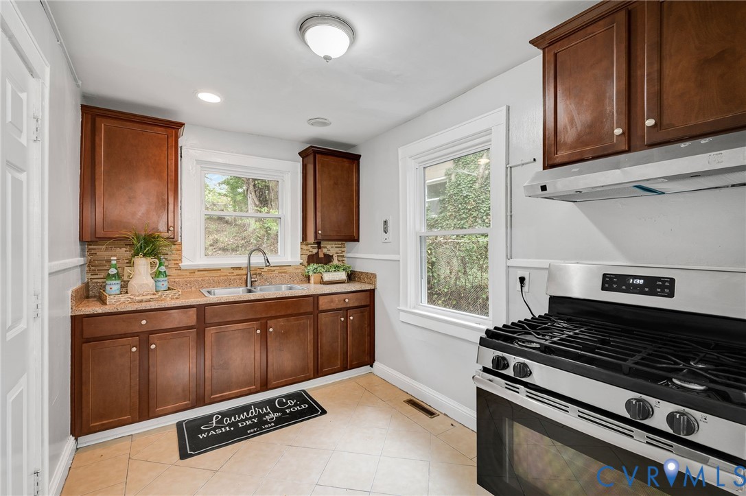 Kitchen featuring stained-wood cabinetry, stone-texture countertops, tile backsplash, stainless steel gas range, and dual-basin stainless steel sink