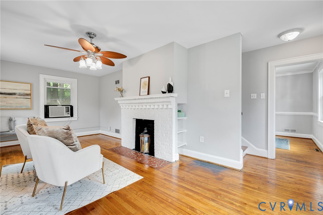 Living room featuring wood-finish flooring, a painted brick fireplace with a decorative mantel, integrated wall shelving, and a ceiling fan with multi-blade design Living room featuring wood-finish flooring, a painted brick fireplace with a decorative mantel, integrated wall shelving, and a ceiling fan with multi-blade design
