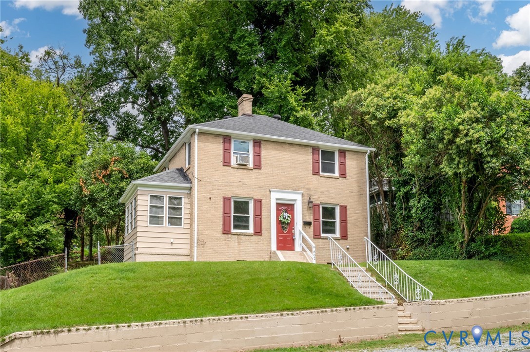 Two-story brick exterior featuring a hipped roof, contrasting window shutters, a front entry with metal railing, and a stone retaining wall Two-story brick exterior featuring a hipped roof, contrasting window shutters, a front entry with metal railing, and a stone retaining wall