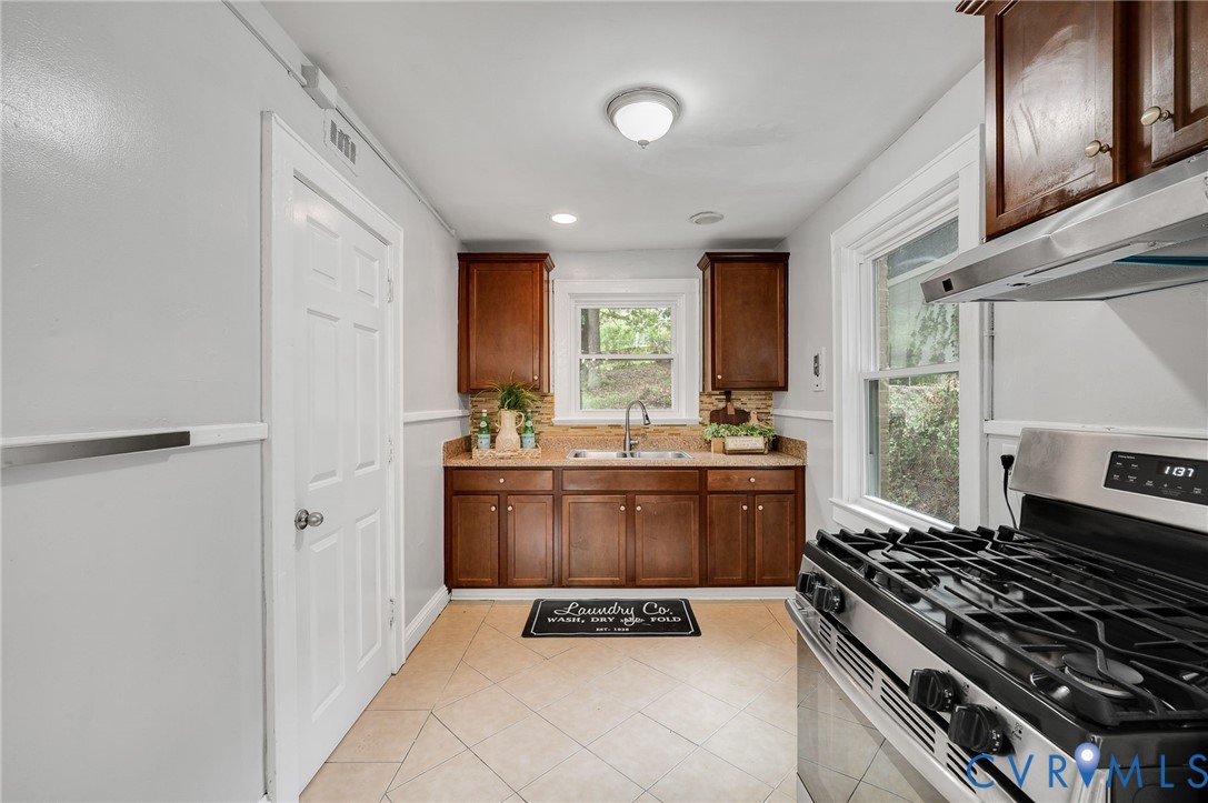 Kitchen featuring stained wood cabinetry, stone-finish countertops, stainless steel gas range with overhead vent hood, tile flooring, and a double-basin sink