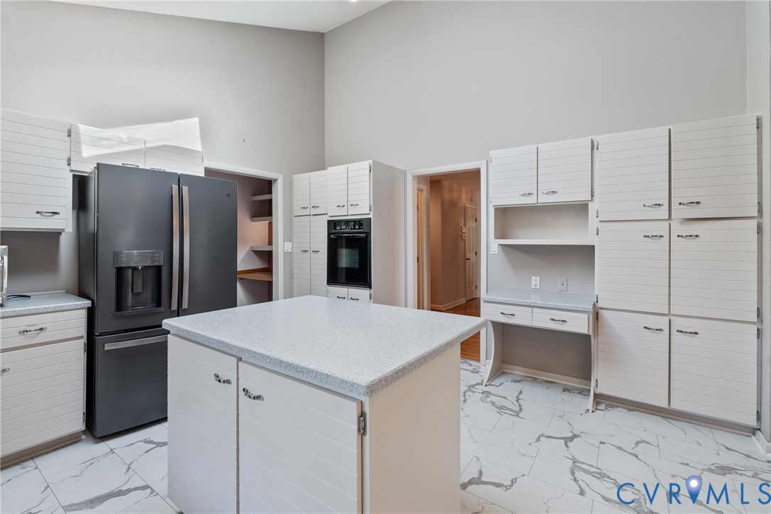 Kitchen featuring white horizontal-paneled cabinetry, a center island with a stone-composite countertop, floor-to-ceiling cabinetry, a built-in wall oven, and light-toned stone-finish flooring with grey veining