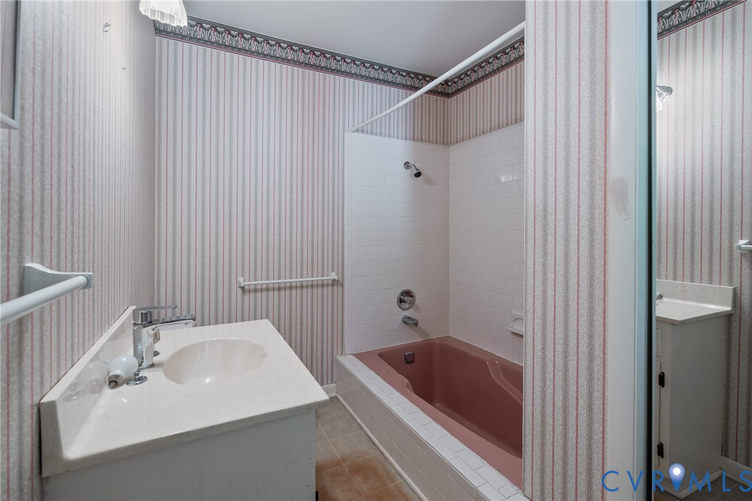 Full bathroom featuring a white vanity cabinet with an integrated sink, polished metal faucet, recessed bathtub with a tiled surround, and decorative wallpaper