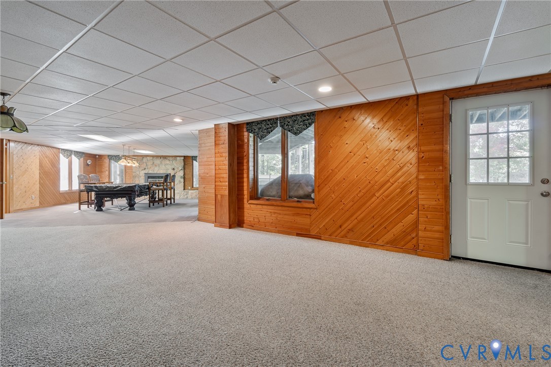Wood-paneled wall accents, stone-faced fireplace, drop ceiling, carpeted flooring, and glass-paneled exterior door