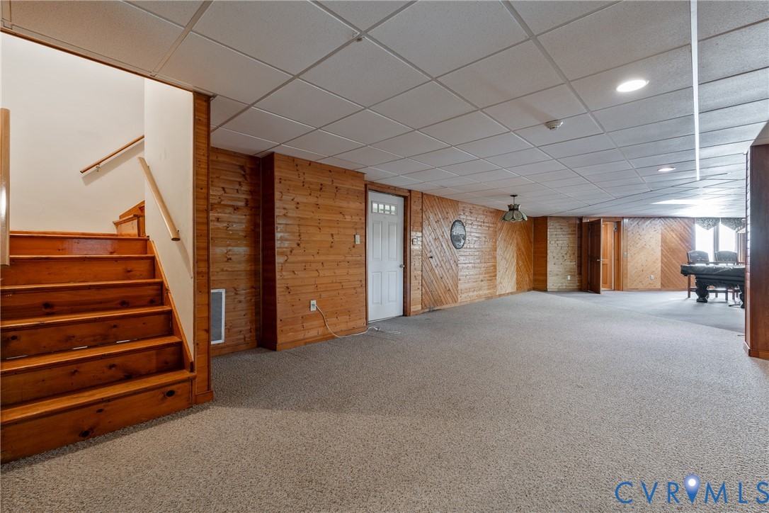Finished room featuring wood-paneled walls, carpeted flooring, a dropped ceiling with recessed lighting, and wooden staircase with handrail
