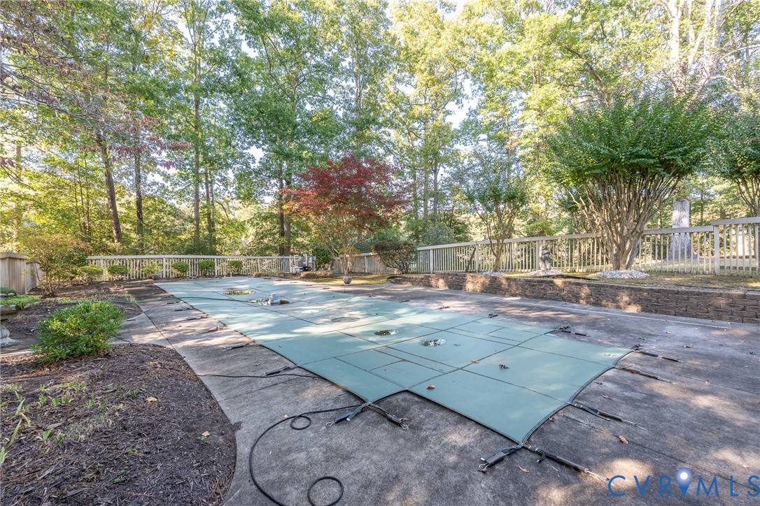 In-ground pool with fitted safety cover, expansive concrete pool deck, stone retaining wall, and wood-slat perimeter fencing surrounded by mature trees