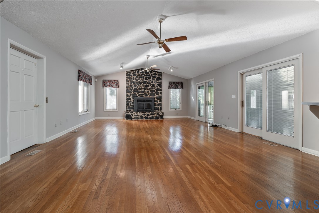 Vaulted ceilings featuring dual ceiling fans, stone-surround fireplace, wood-finish flooring, and multiple glass-paneled exterior doors